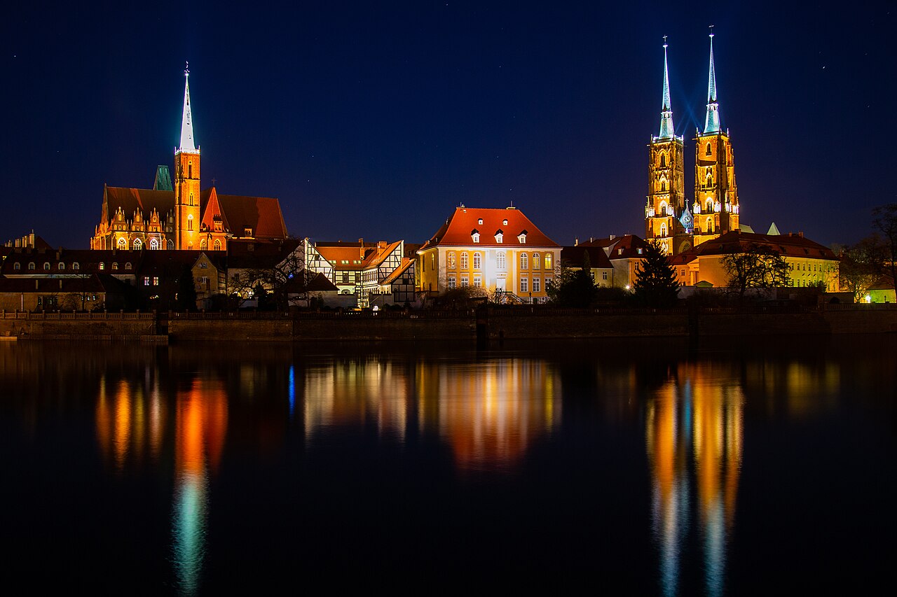 Vista noturna de Ostrów Tumski à margem do rio, com igrejas iluminadas, torres, árvores sem folhas e reflexos dourados na água escura.