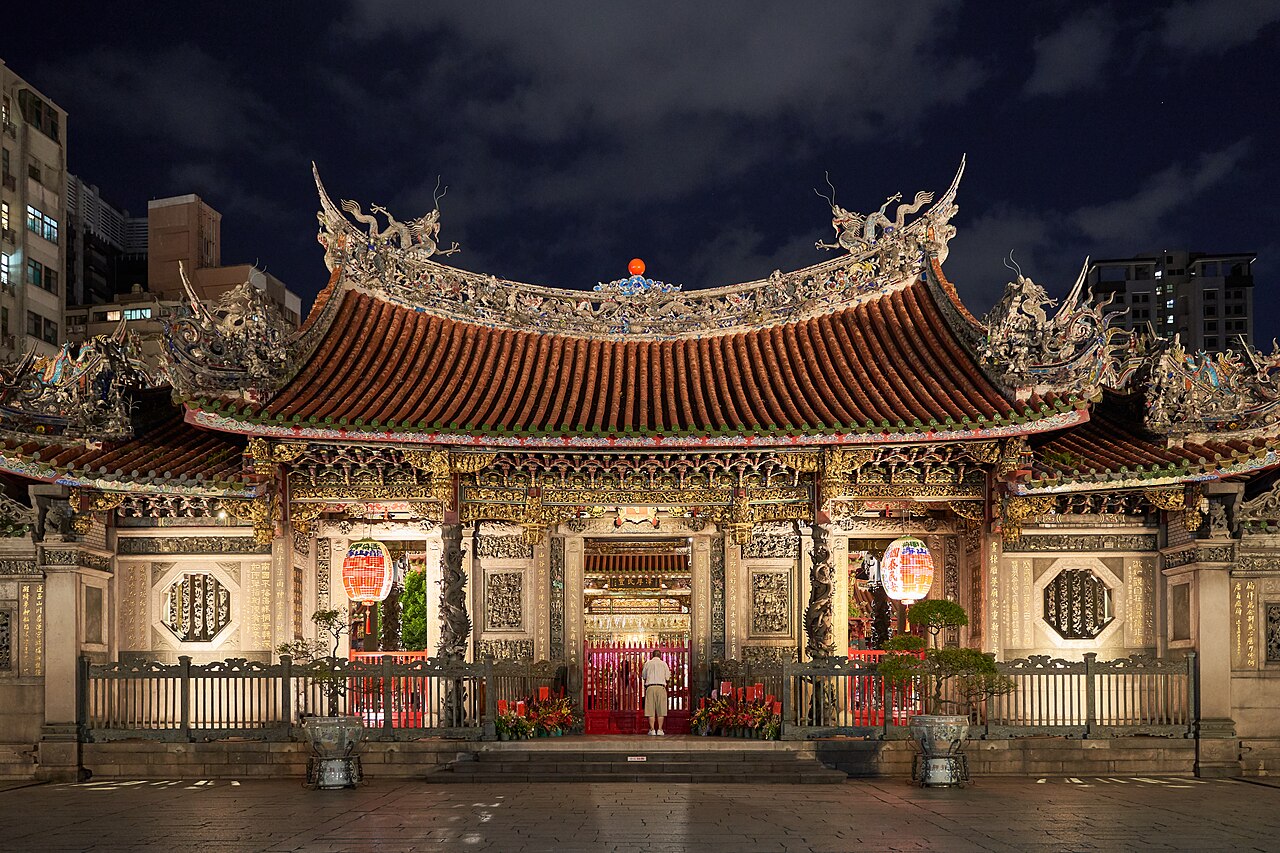 Fachada noturna iluminada do Templo Longshan, com telhado de pontas curvas, dragões, colunas esculpidas e lanternas.