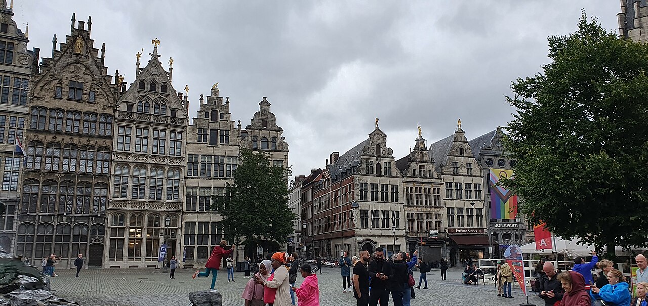 Praça da Grote Markt com pedestres, fachadas estreitas de frontões ornamentados e mesas à direita, sob céu nublado.
