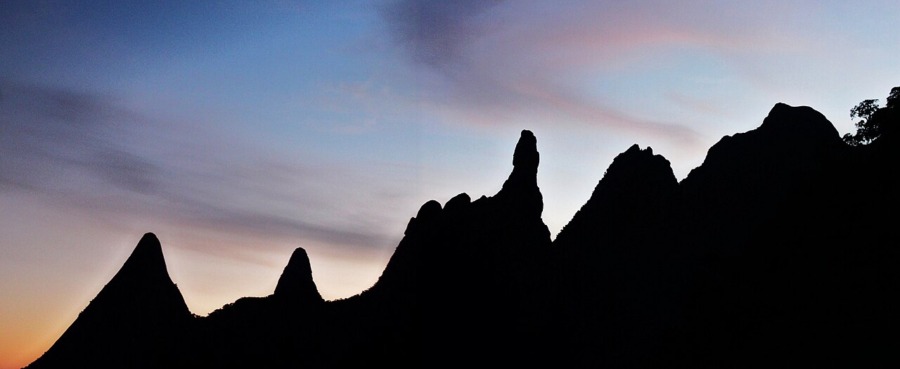 Cadeia de montanhas em silhueta escura, com picos pontiagudos, diante de um céu azul, rosado e alaranjado.