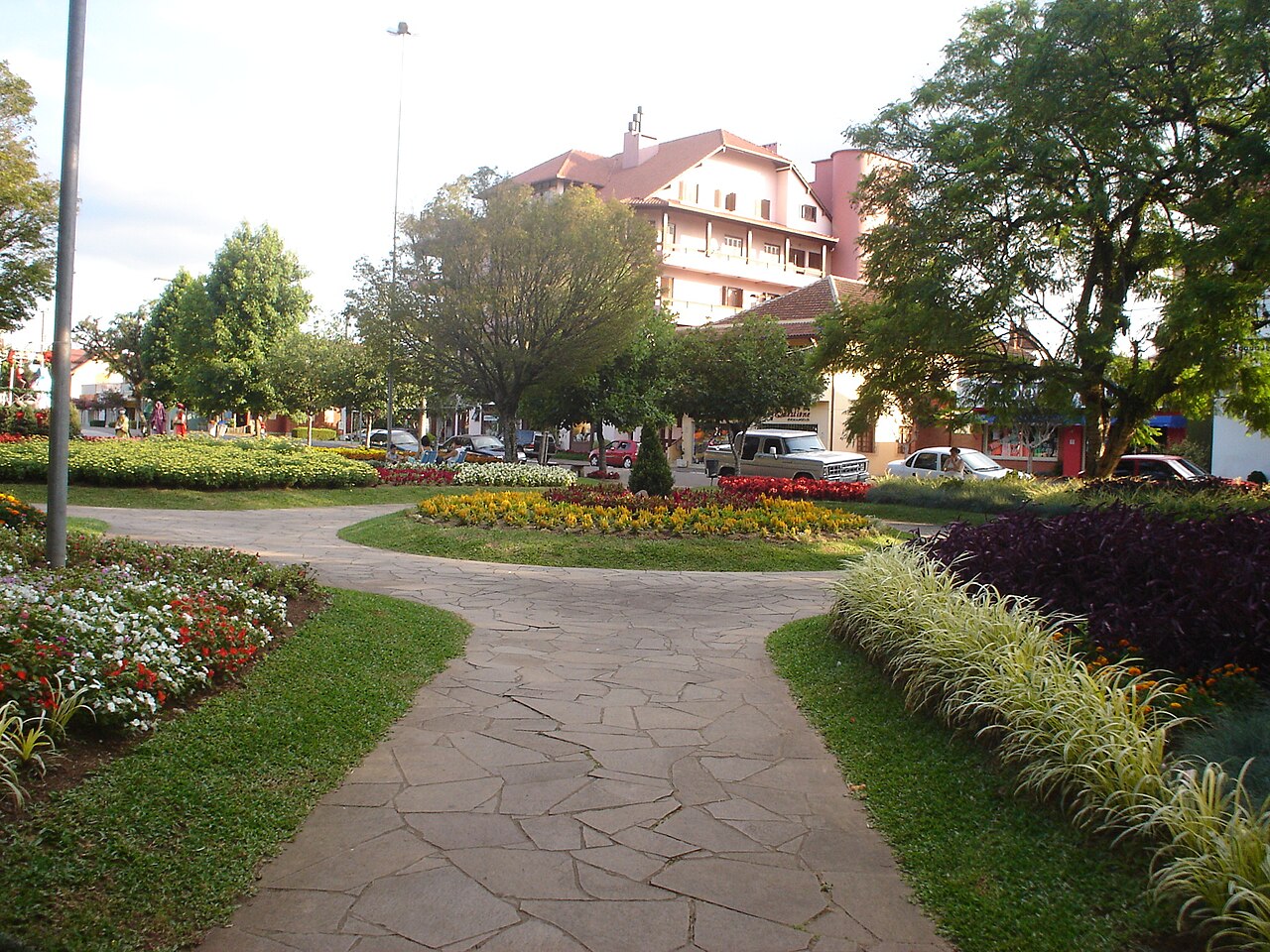 Praça das Flores com caminhos de pedra, gramados, canteiros coloridos, árvores e edifícios ao fundo.