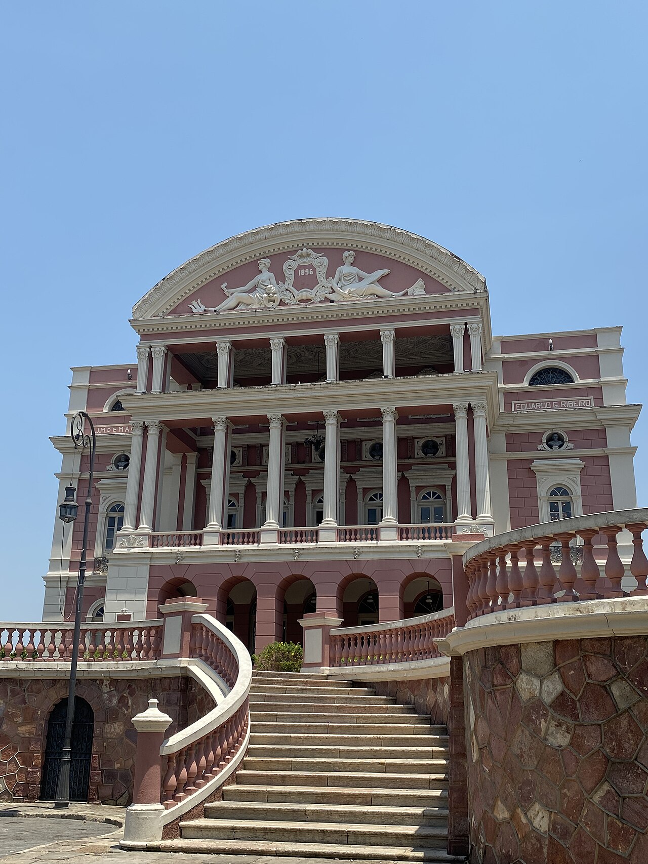 Fachada rosa do Teatro Amazonas vista de baixo, com colunas claras, frontão com esculturas, escadaria e balaustradas sob céu azul.