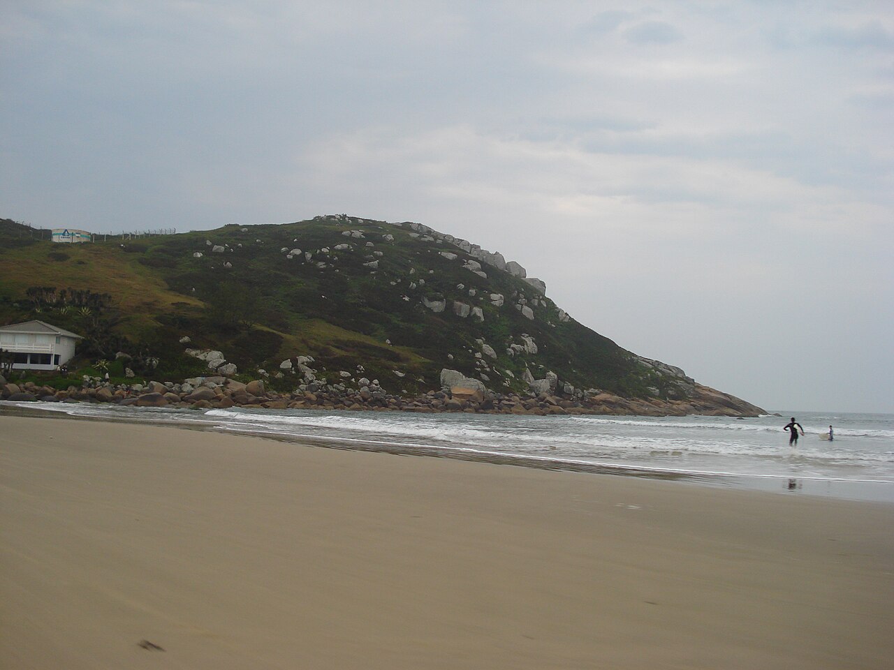 Praia de areia larga com pequenas ondas, duas pessoas na água e um morro com vegetação e grandes rochas sob céu encoberto.