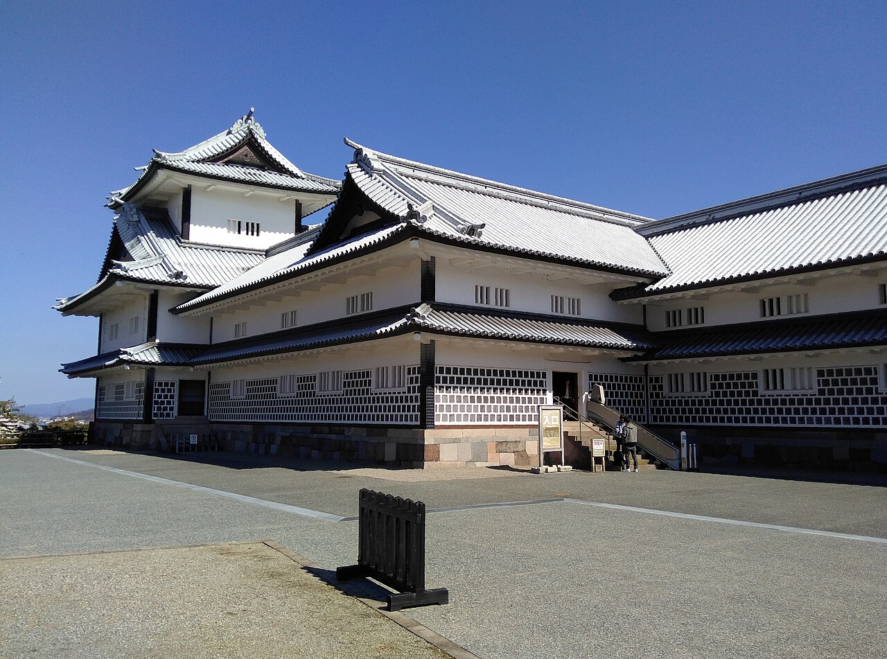 Torre Hishi Yagura e conjunto de paredes brancas do Castelo de Kanazawa, com telhados claros, base de pedra e pátio sob céu azul.
