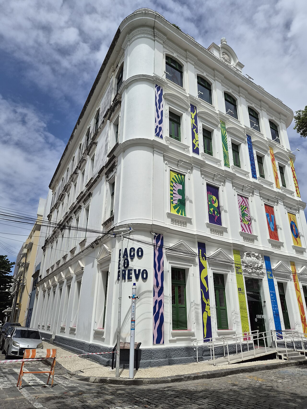 Fachada branca de esquina do Paço do Frevo, com janelas verdes, faixas multicoloridas e inscrição com o nome do museu.