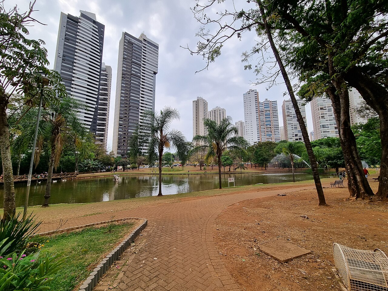 Parque Flamboyant com lago, caminho curvo de tijolos, bancos, palmeiras, pessoas e torres residenciais ao fundo.