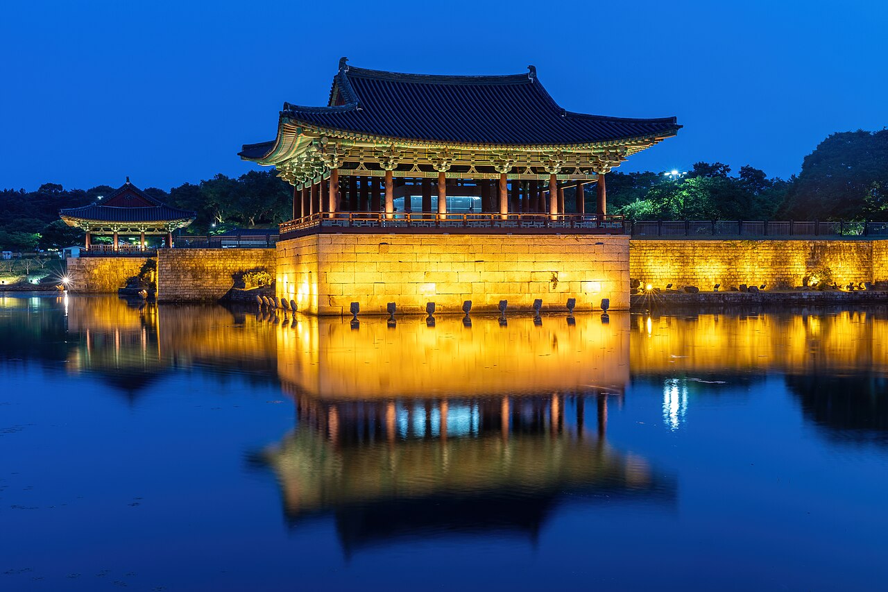 Pavilhões de Donggung iluminados junto ao lago Wolji, com reflexos dourados na água e céu azul ao anoitecer.