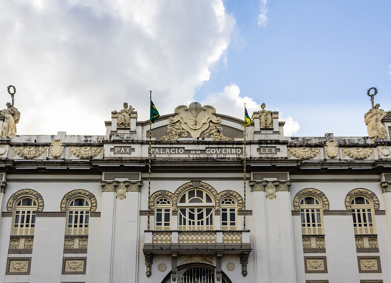Trecho da fachada clara do Palácio Olímpio Campos, com relevos ornamentais, janelas arqueadas, sacada, bandeiras e estátuas no topo.