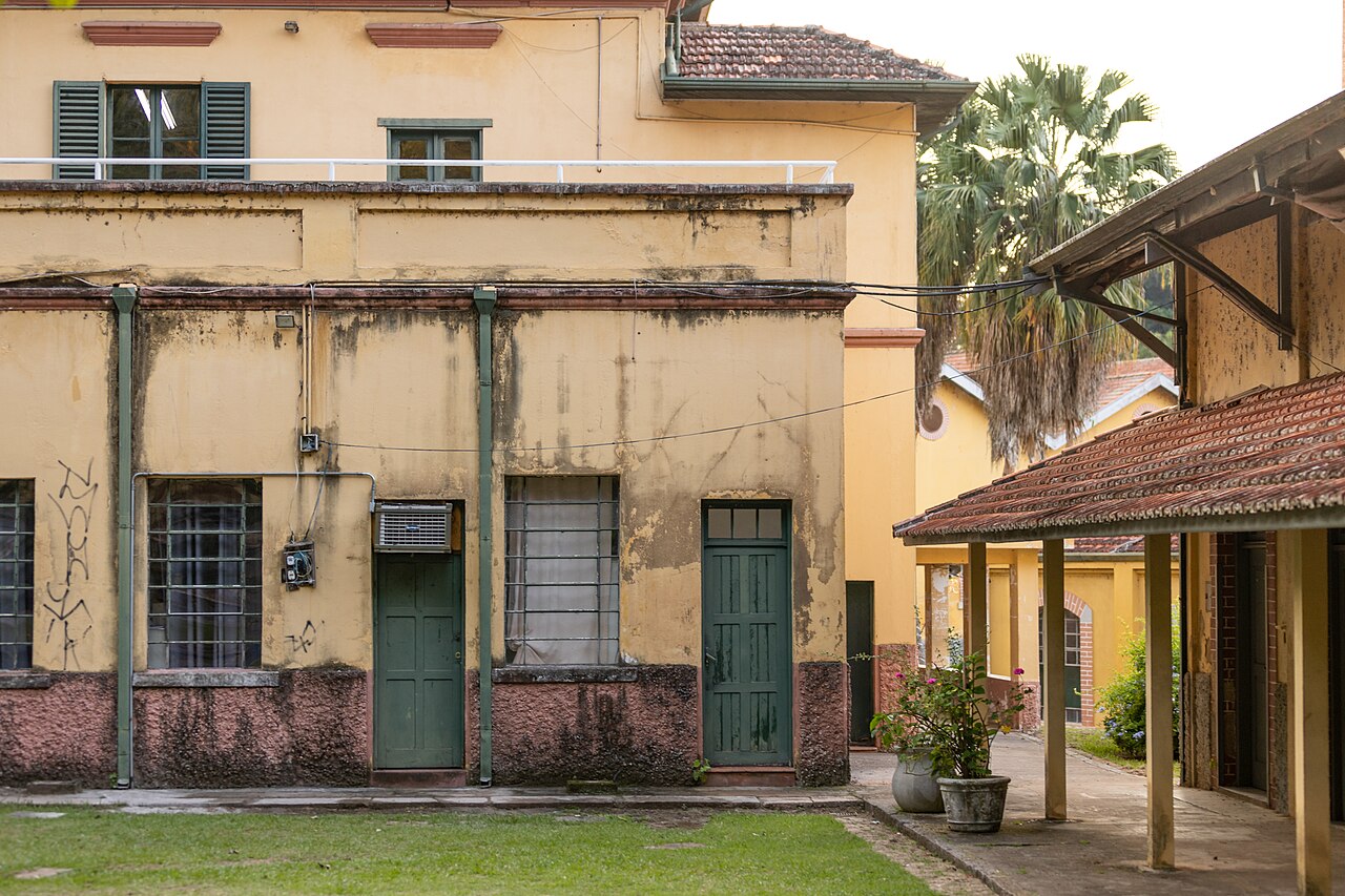 Pátio gramado da Brasital com edifícios amarelos, portas e esquadrias verdes, telhados cerâmicos e passagem coberta à direita.