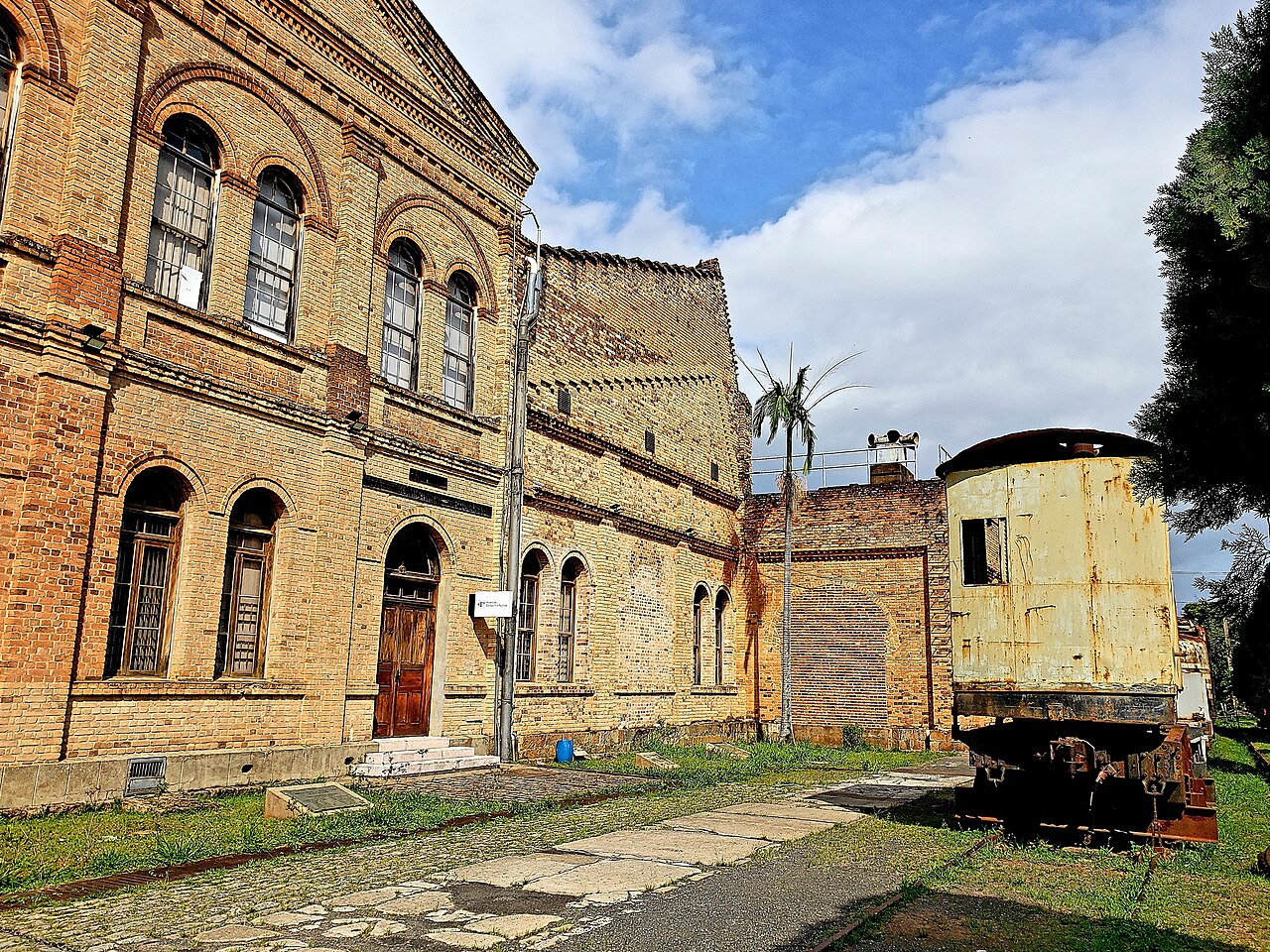 Edifícios de tijolos aparentes com portas e janelas em arco e um veículo ferroviário antigo sobre trilhos à direita.