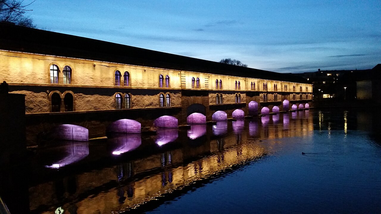 Barrage Vauban sobre a água ao entardecer, com fachada iluminada em amarelo e luz roxa nos arcos refletida no canal.