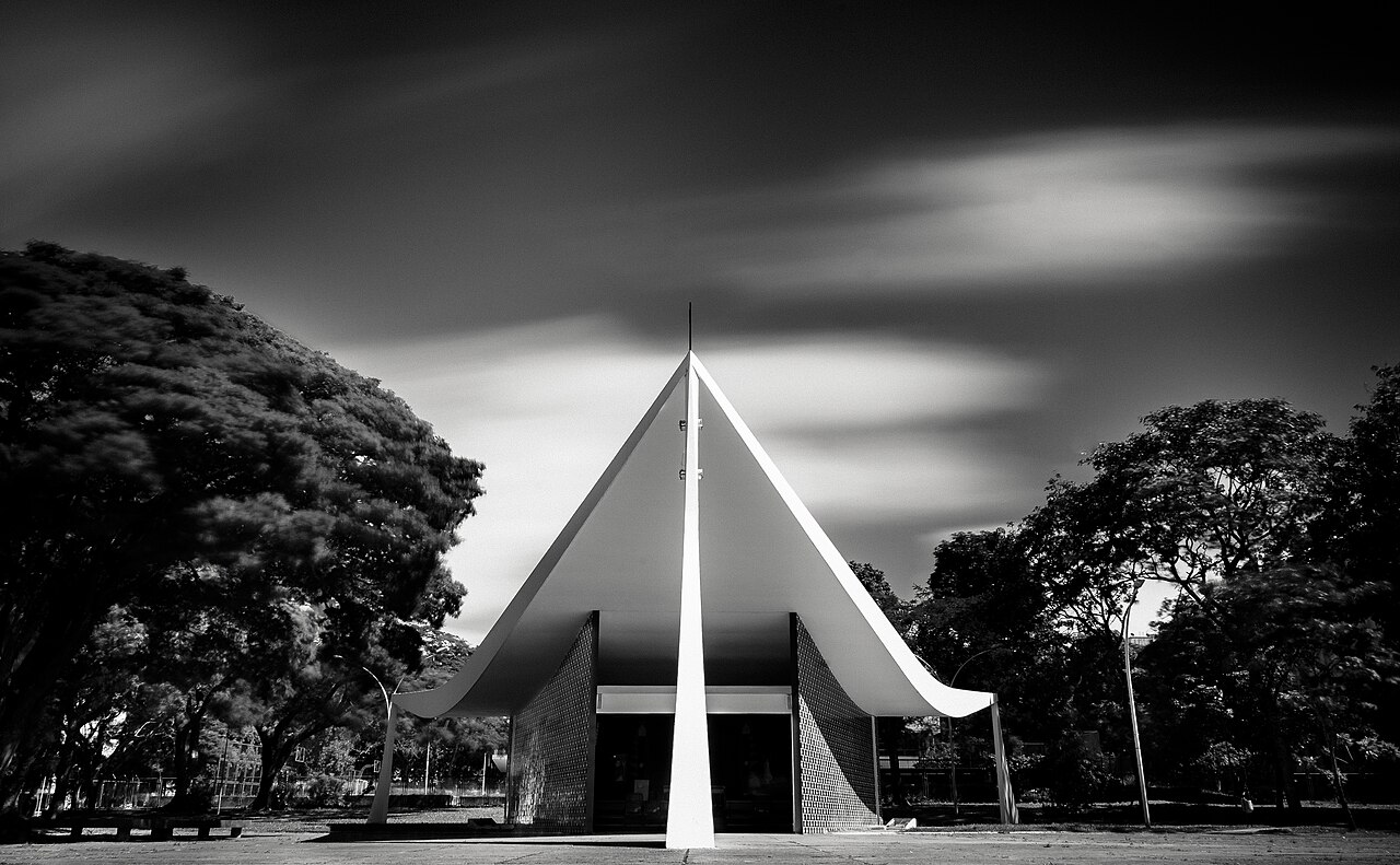 Fotografia em preto e branco da Igrejinha Nossa Senhora de Fátima, com cobertura triangular curva, paredes de azulejos e árvores ao redor.