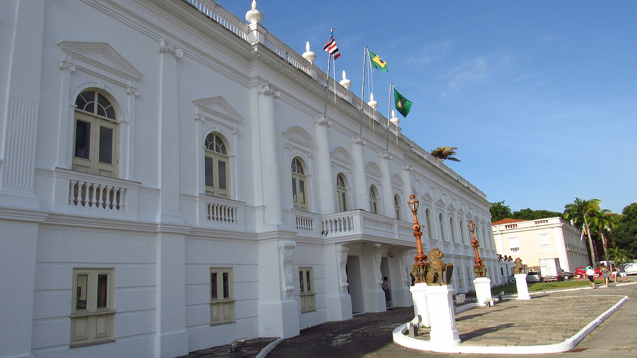 Fachada branca do Palácio dos Leões, com janelas altas, balaustradas, bandeiras e esculturas de leões sobre pedestais.