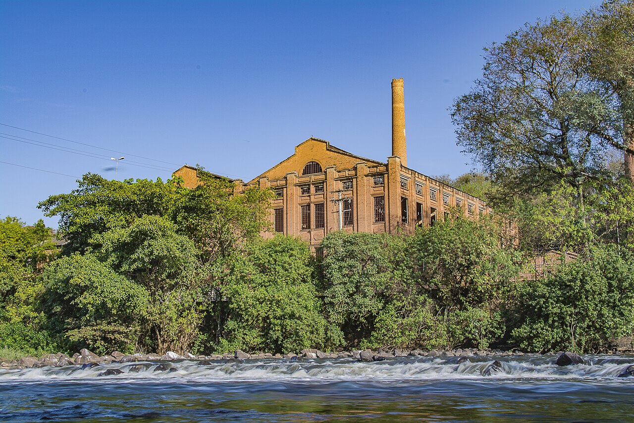 Edifício industrial de tijolos com grandes janelas e chaminé atrás de vegetação, com rio, pedras e corredeiras em primeiro plano.