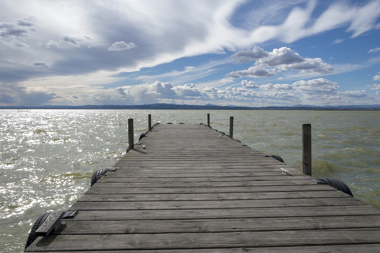 Passarela de madeira sobre a água da Albufera, com postes, amarras e pneus nas laterais, sem pessoas ou embarcações visíveis.