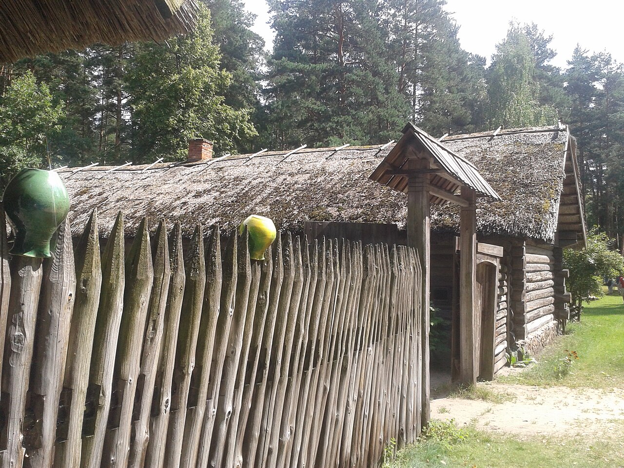 Construção de troncos no conjunto de Latgale do Museu Etnográfico, com cobertura de palha, cerca de madeira, recipientes de cerâmica e árvores ao redor.