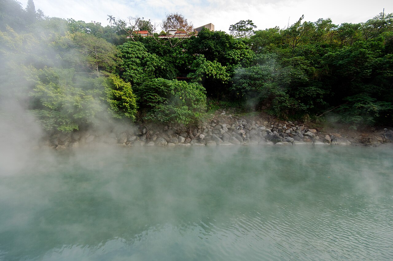 Água verde-azulada de Thermal Valley com vapor sobre a superfície, margem rochosa e vegetação densa.