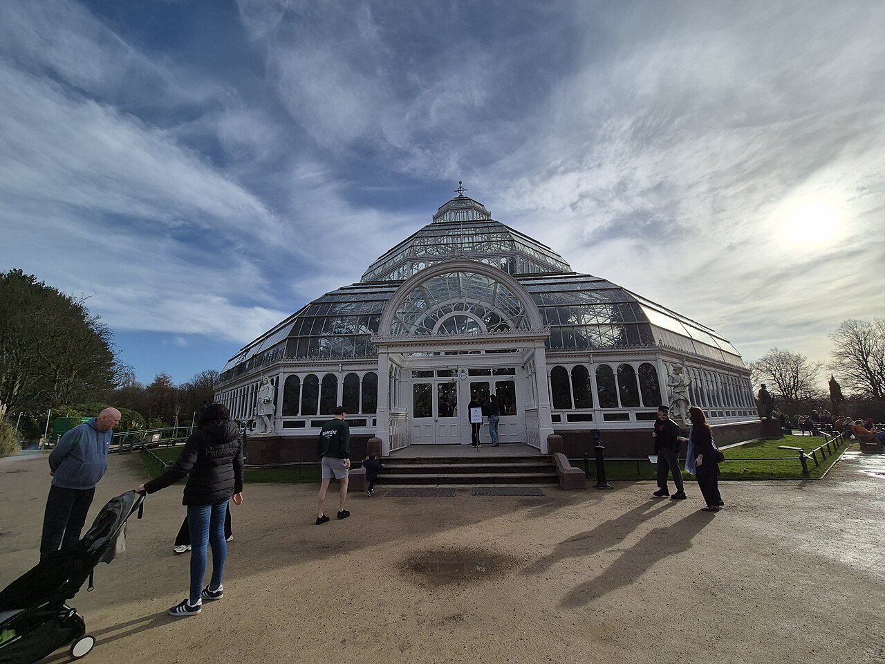 Fachada da Palm House, uma estufa de vidro e metal com entrada central em arco, estátuas, visitantes, caminhos e gramados ao redor.
