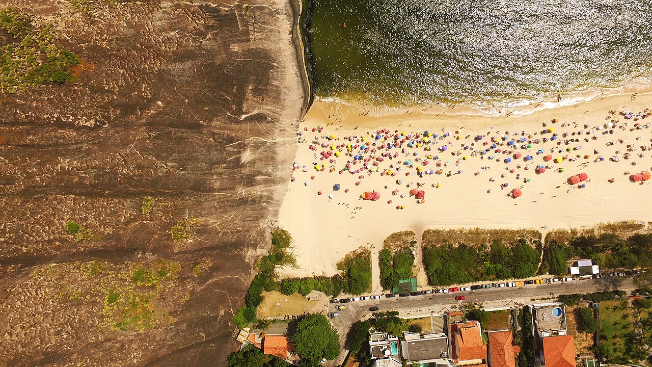Vista aérea de Itacoatiara com formação rochosa à esquerda, areia com banhistas e guarda-sóis, mar e uma rua junto às casas.