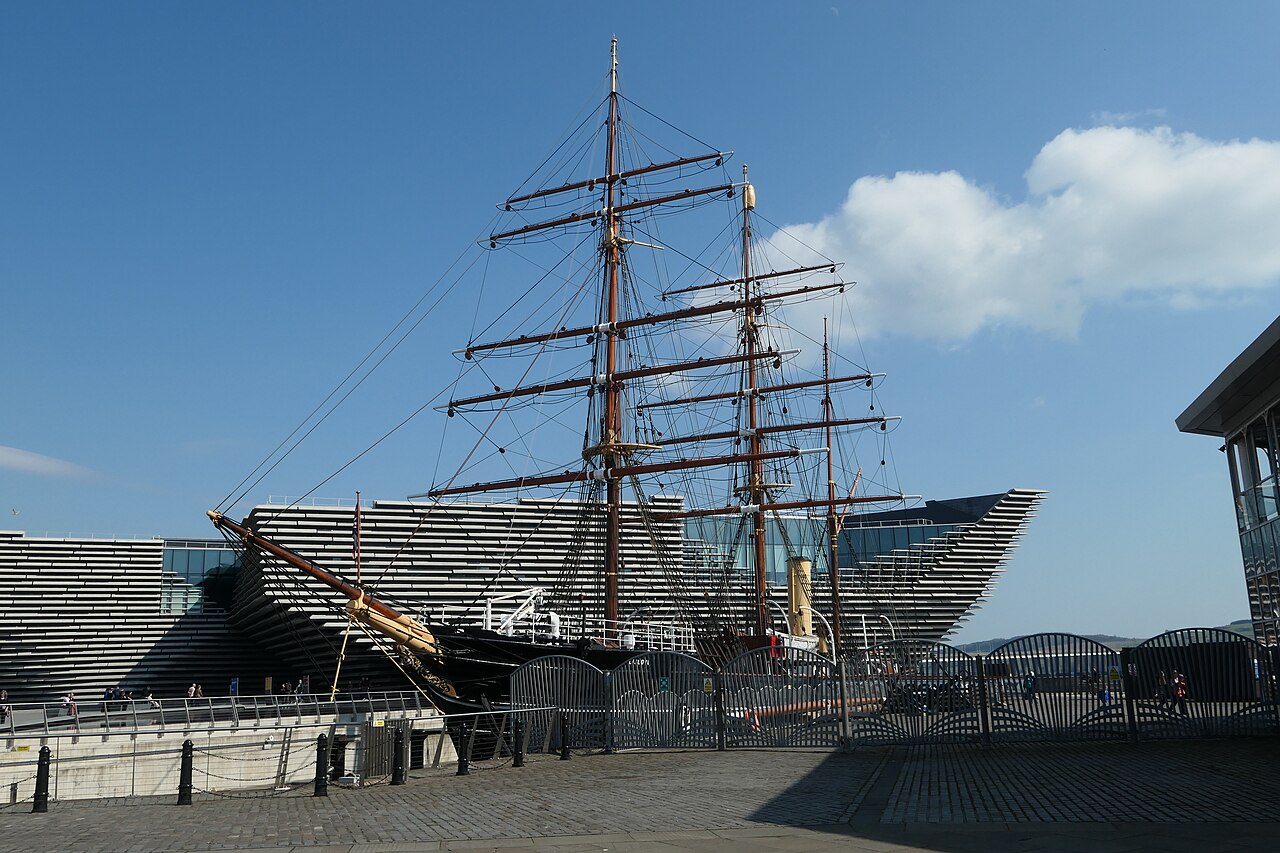 Navio histórico de três mastros e casco escuro junto ao cais, com cordames, chaminé clara e o edifício do V&A ao fundo.