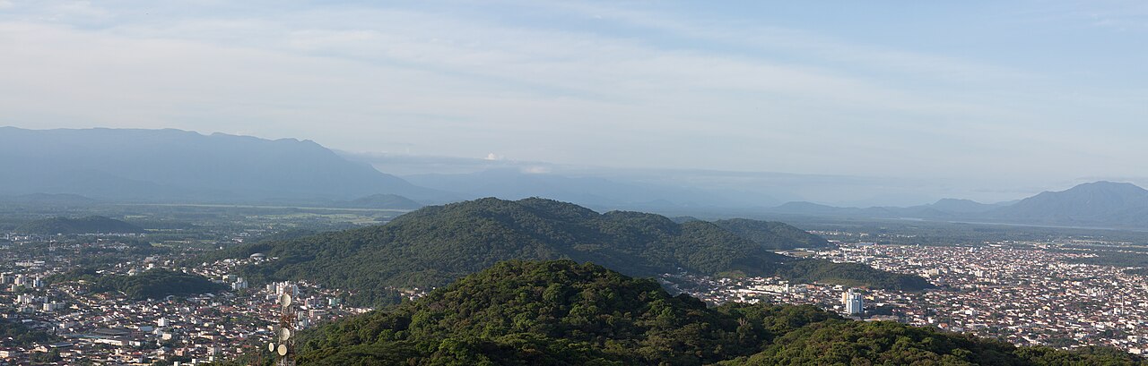 Panorama elevado de áreas urbanizadas, morros verdes e serras azuladas, com uma torre de antenas em primeiro plano.