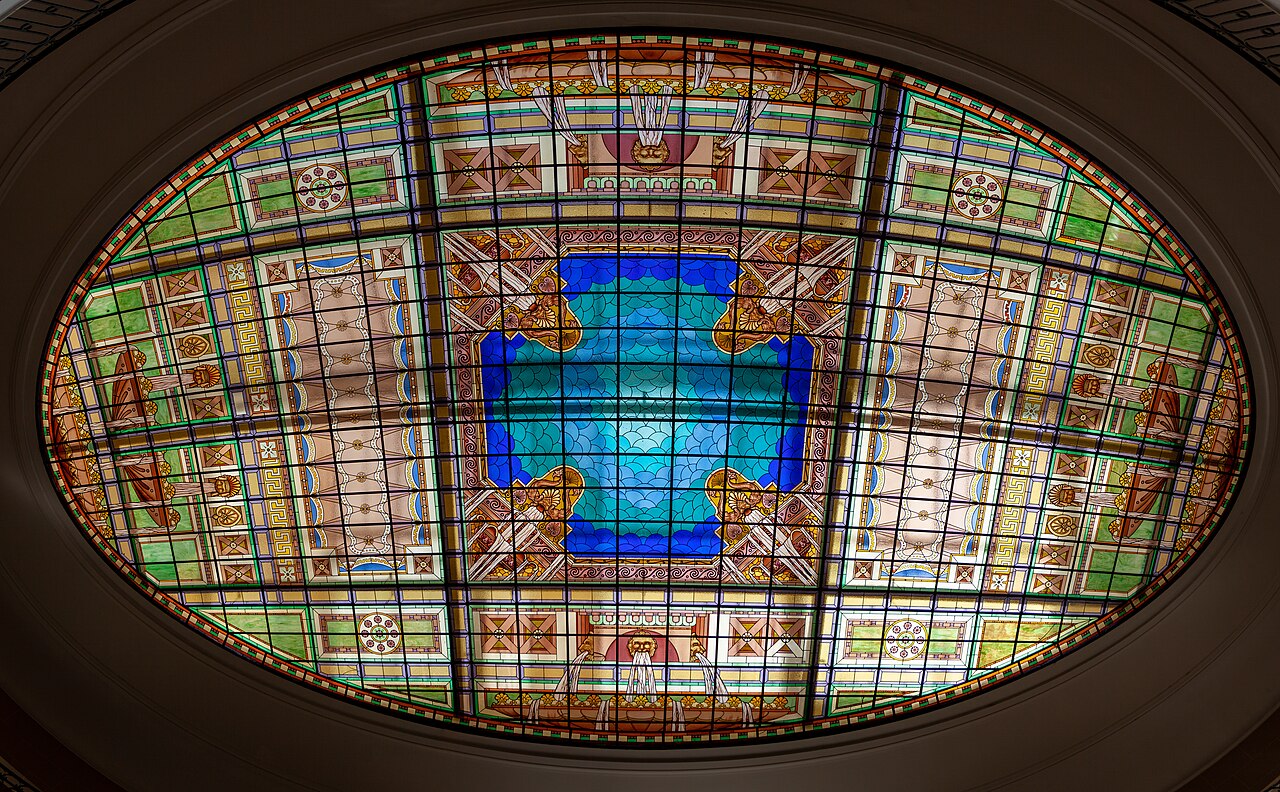Vitral oval visto de baixo, com centro azul e turquesa, faixas verdes e motivos geométricos e florais em uma moldura clara.