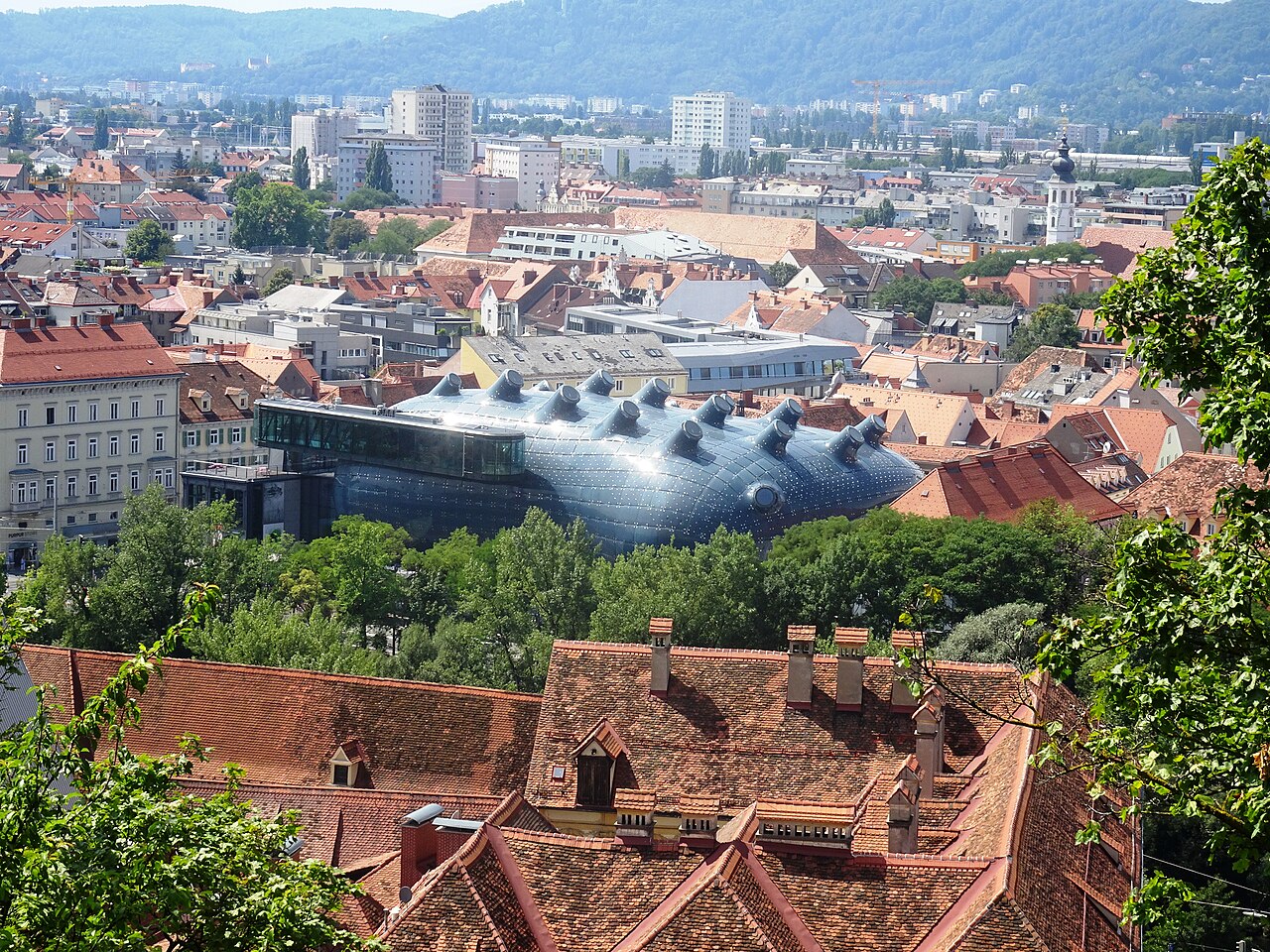 Vista elevada do Kunsthaus, com volume escuro arredondado e protuberâncias tubulares, entre telhados vermelhos e edifícios de Graz.