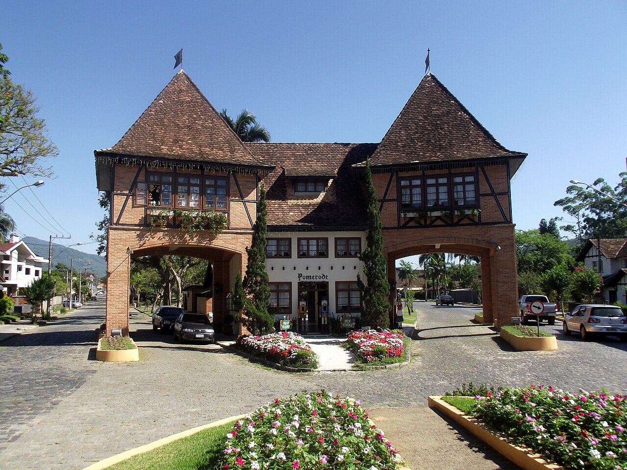 Portal com dois arcos, tijolos aparentes, madeira em estilo enxaimel e inscrição Pomerode, cercado por canteiros e ruas.