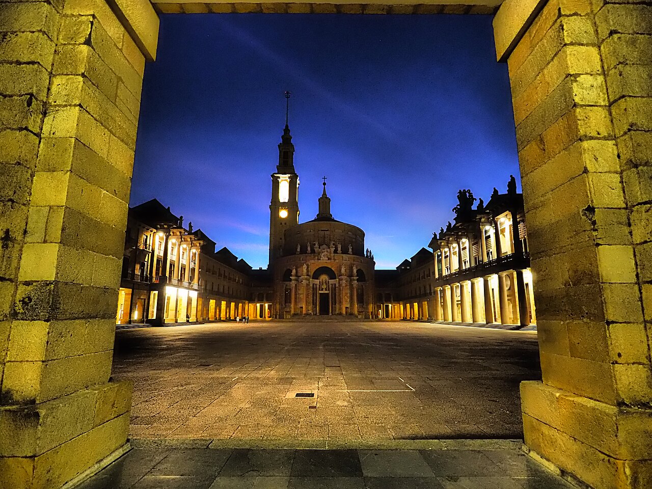 Pátio amplo visto através de um portal de pedra, com torre, edifício arredondado e colunatas iluminadas sob céu azul-escuro.