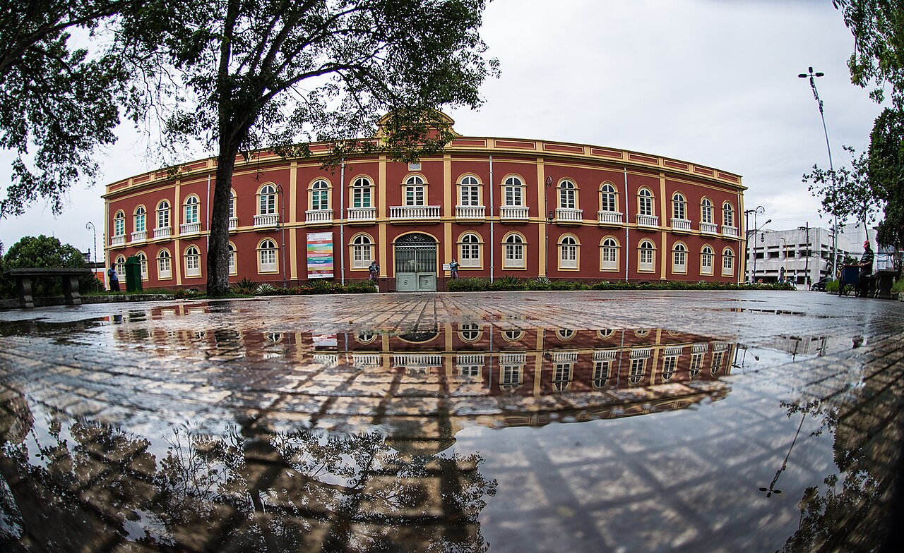 Fachada avermelhada de dois pavimentos do Palacete Provincial, com detalhes amarelos, árvores ao redor e reflexos em poças na praça.
