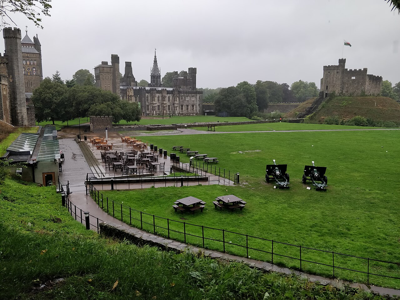 Pátio de Cardiff Castle com gramado, mesas, bancos e dois canhões, edifícios de pedra ao fundo e fortificação sobre uma elevação à direita.