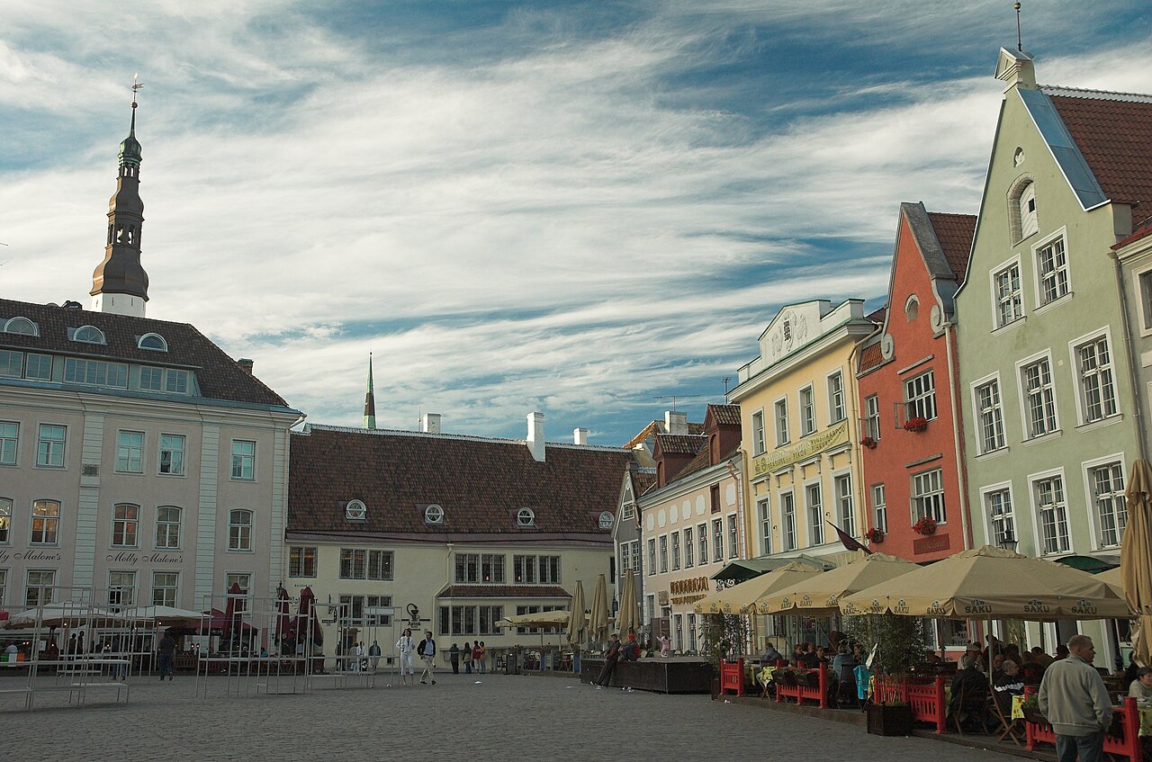 Praça da Prefeitura de Tallinn com piso de pedra, fachadas claras e coloridas, torre à esquerda e mesas sob guarda-sóis à direita.