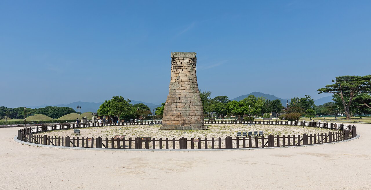 Torre de pedra de Cheomseongdae com abertura quadrada, cercada por caminhos, gramados e árvores sob céu azul.