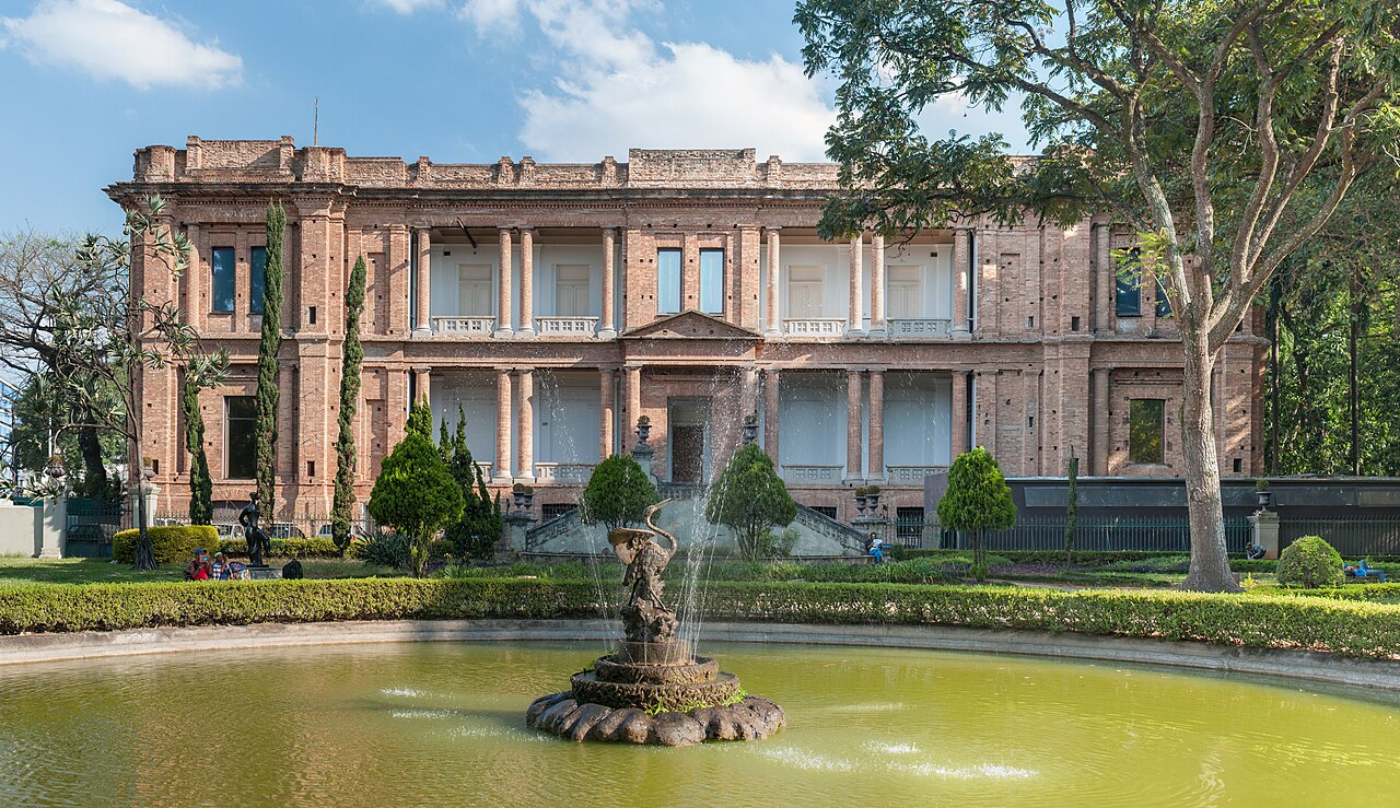 Fachada de tijolos aparentes da Pinacoteca, com colunas e grandes janelas, diante de jardins e de um lago com chafariz e escultura.