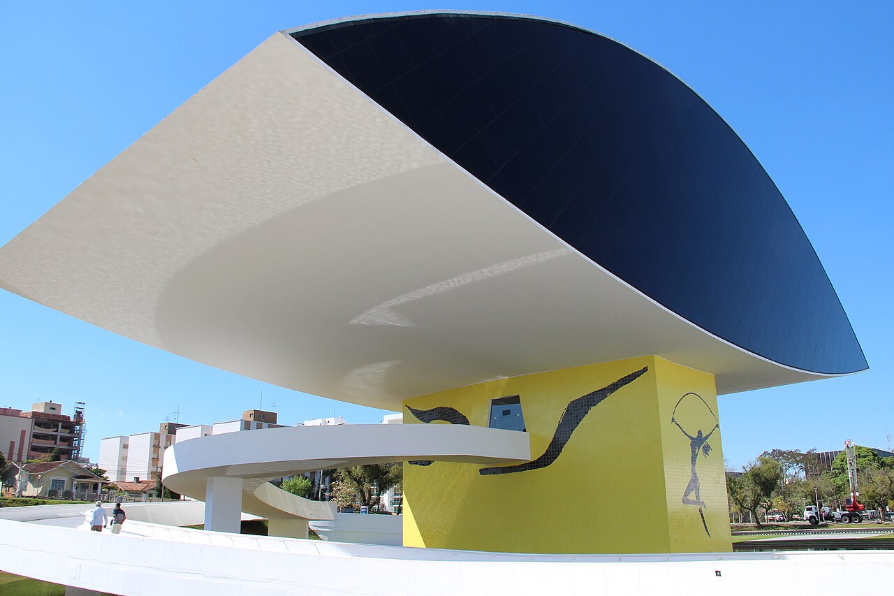 Estrutura elevada em forma de olho do Museu Oscar Niemeyer, com apoio amarelo e rampa branca curva sob céu azul.
