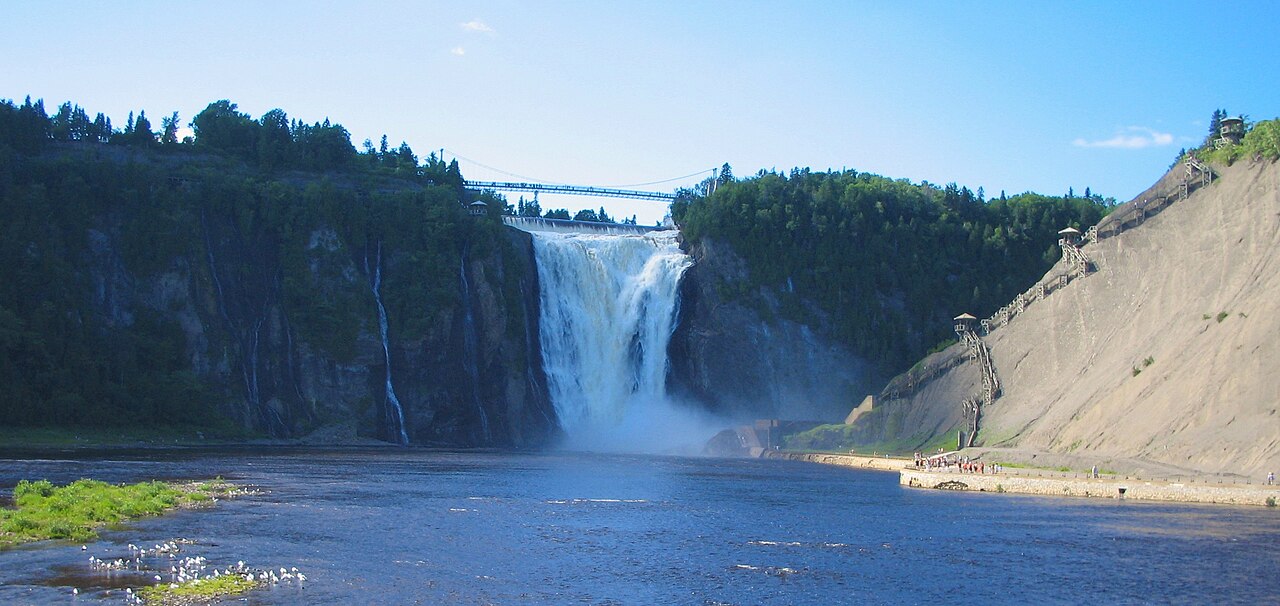 Queda Montmorency entre paredões rochosos, com ponte suspensa acima da água, escadarias e mirantes na encosta direita e visitantes na parte inferior.