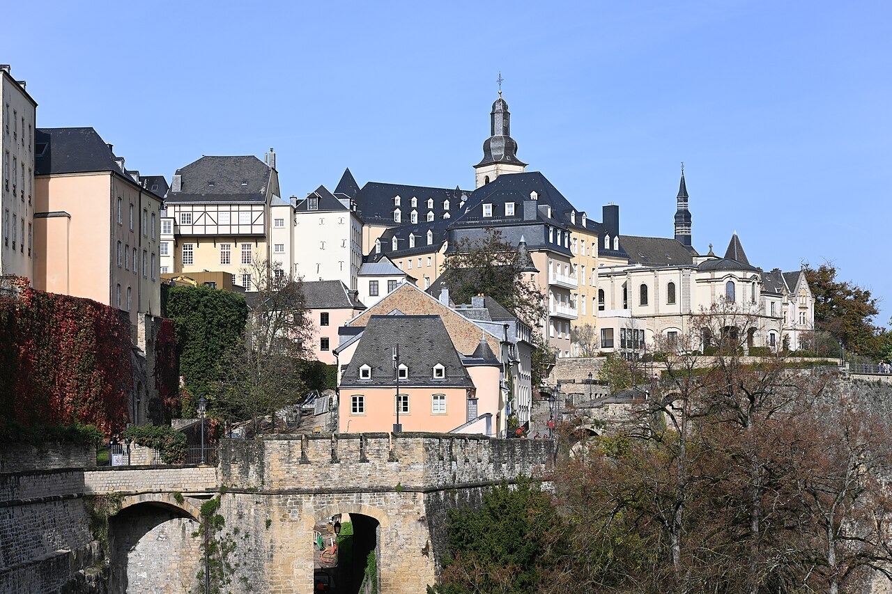 Muralhas de pedra com arcos, edifícios claros de telhados escuros, torres e árvores na área da Corniche, sob céu azul.