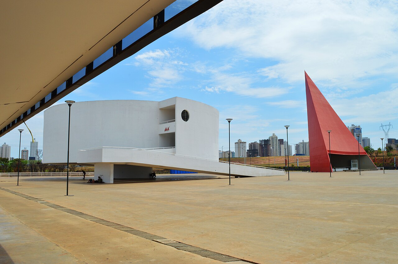 Esplanada do Centro Cultural Oscar Niemeyer com edifício branco curvo, rampa e estrutura vermelha triangular sob céu azul.
