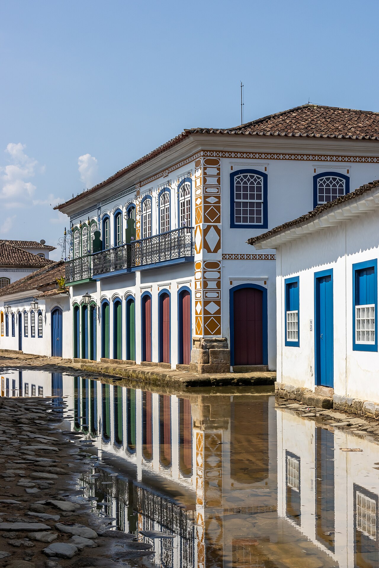 Rua de pedras parcialmente coberta por água, com reflexos de casarões brancos, portas coloridas e sacadas de ferro em Paraty.