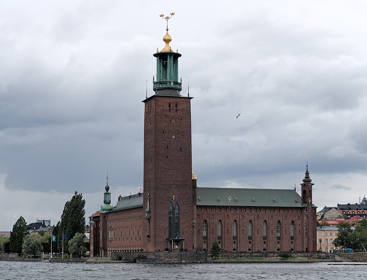 Exterior de tijolos vermelhos do Stadshuset à beira d’água, com torre alta, telhados verdes e três coroas no topo.