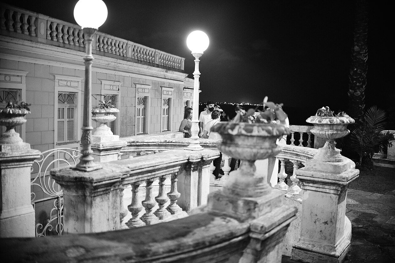 Fotografia noturna em preto e branco de parte da fachada do Hotel Globo, com venezianas, balaustradas, vasos e postes iluminados.