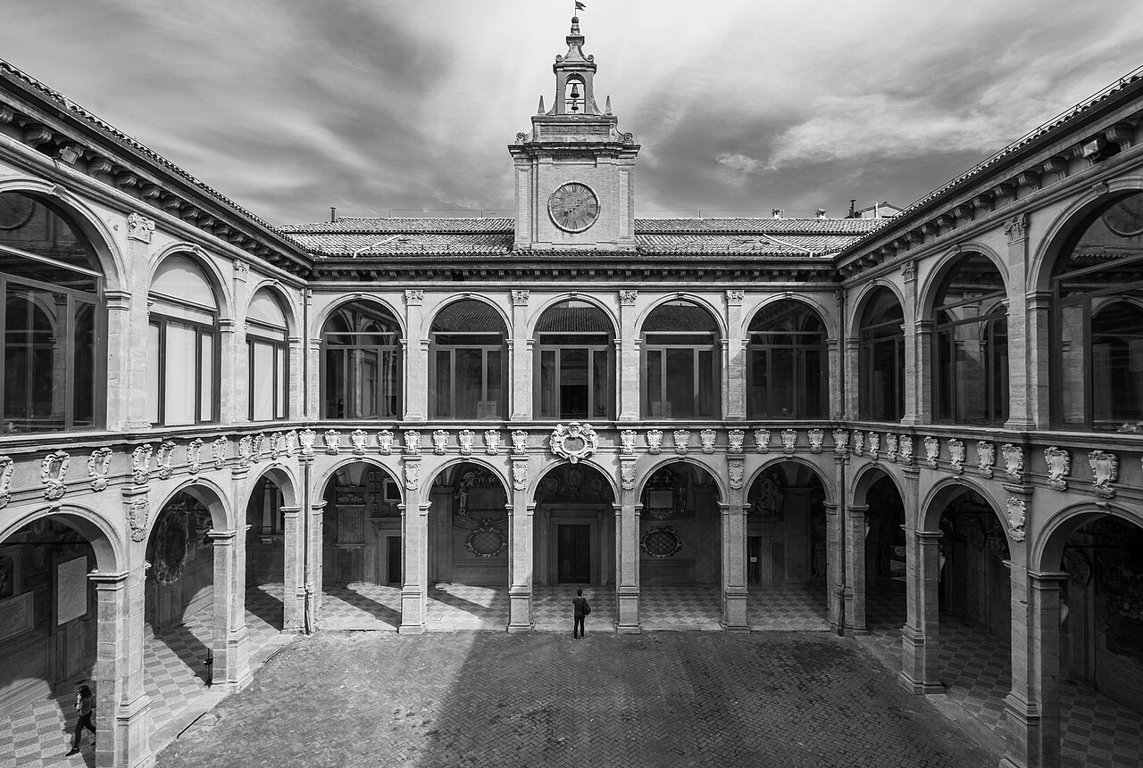 Fotografia em preto e branco do pátio do Archiginnasio, com galerias de arcos em dois níveis, brasões e uma torre com relógio e sino.