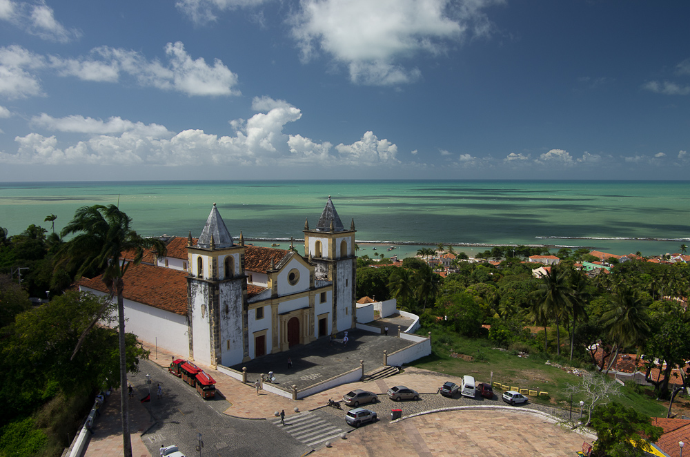 Vista elevada da Catedral da Sé, branca com detalhes amarelos e duas torres, diante de um largo com escadarias, pessoas e veículos; o mar aparece ao fundo.