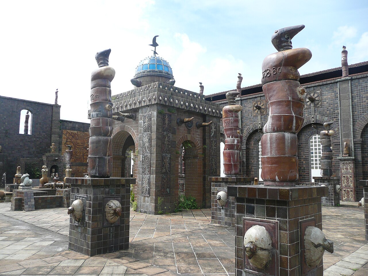 Pátio da Oficina Francisco Brennand com revestimentos cerâmicos, esculturas verticais em tons terrosos, arcos e uma cúpula azul.