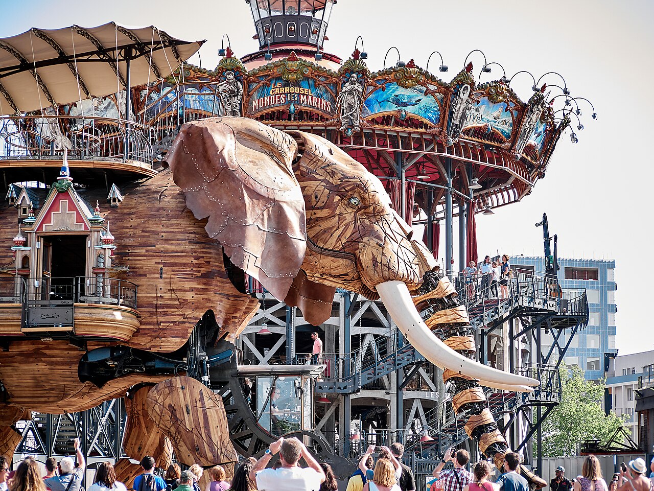 Grande elefante mecânico revestido de madeira, com mecanismos expostos, visitantes e plataformas; o Carrousel des Mondes Marins aparece ao fundo.
