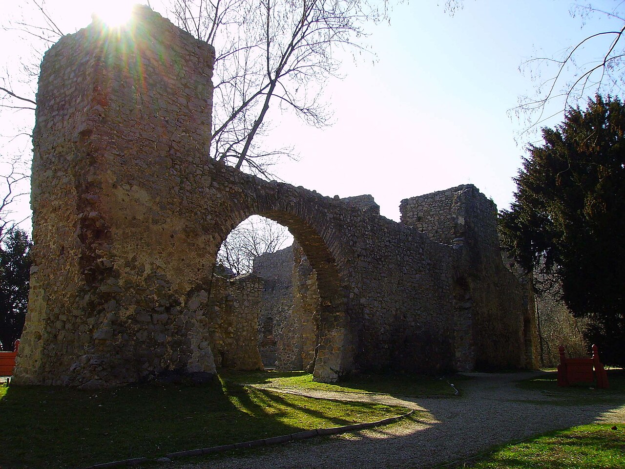 Ruínas de pedra de Tettye com paredes altas e um grande arco, cercadas por gramado, caminhos, bancos e árvores.