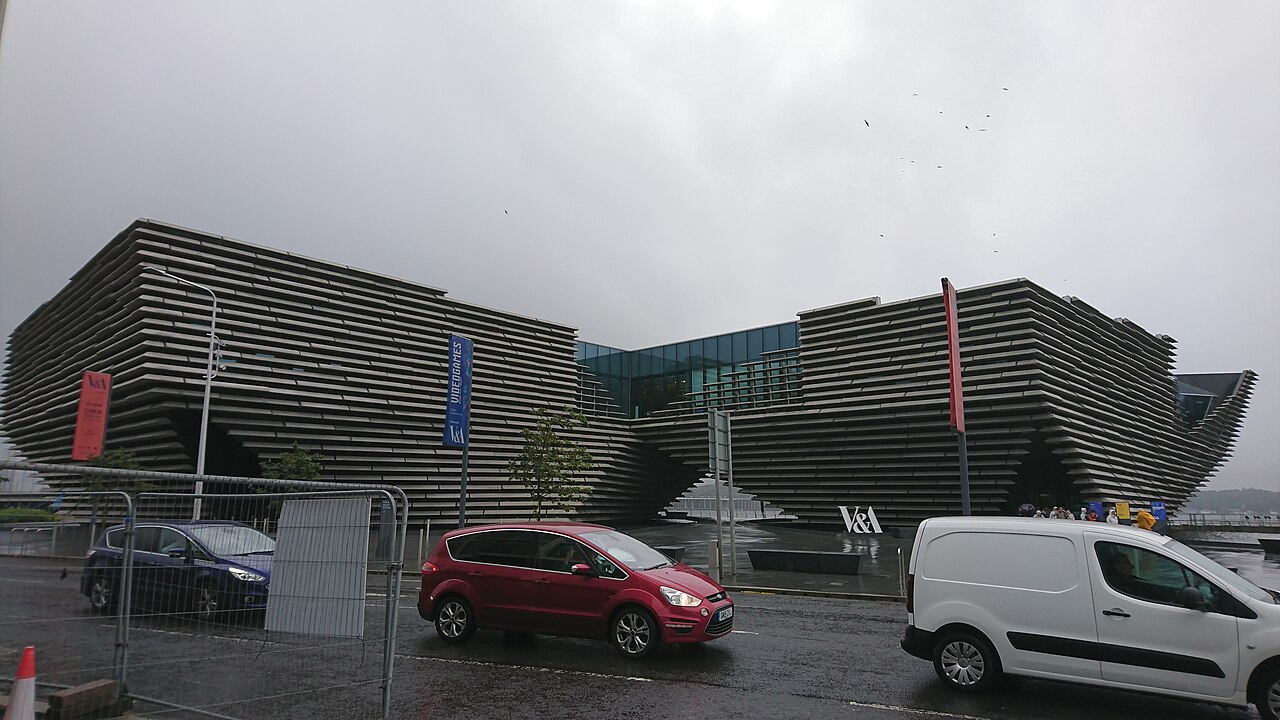 Fachada angular do V&A Dundee com faixas horizontais, área central envidraçada e letreiro, diante de pavimento molhado, veículos e grades de obra.