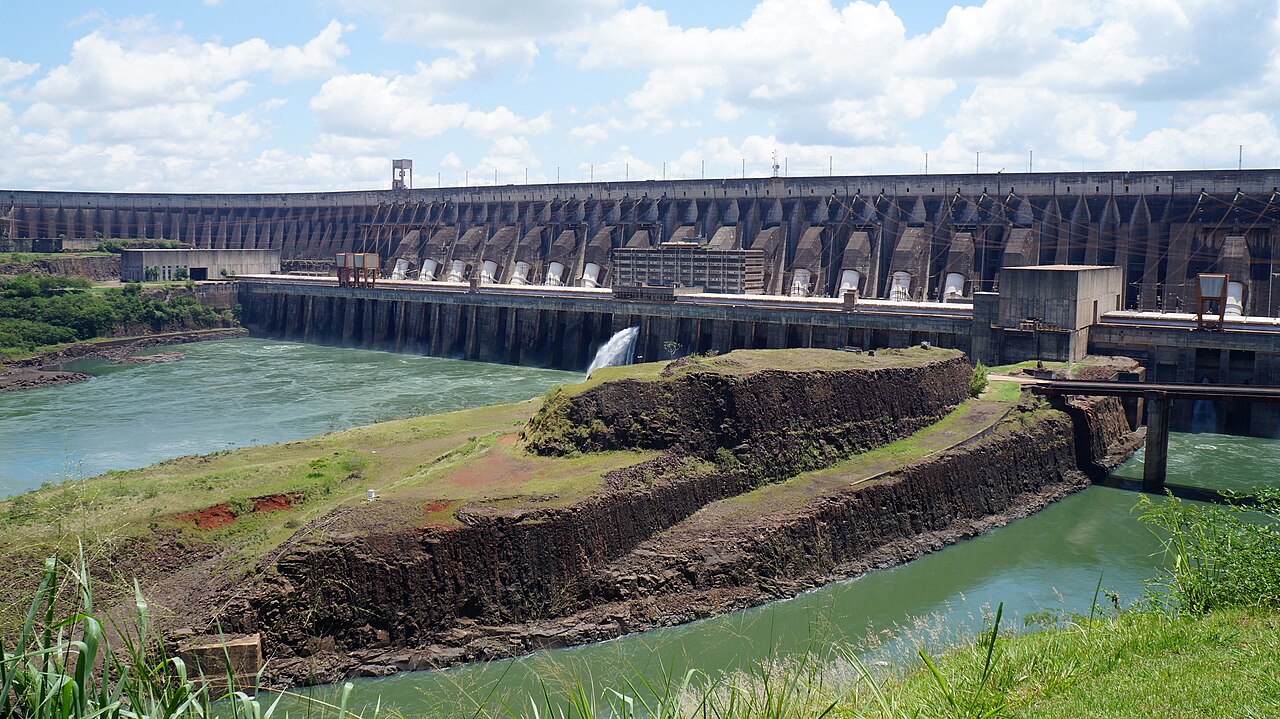 Barragem de concreto de Itaipu, com instalações na base, água esverdeada e taludes rochosos.