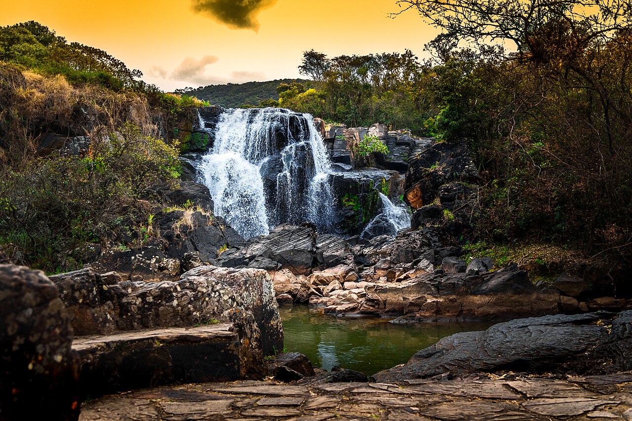 Quedas de água sobre rochas escuras, com pequeno poço esverdeado, lajes em primeiro plano, vegetação e céu amarelado.