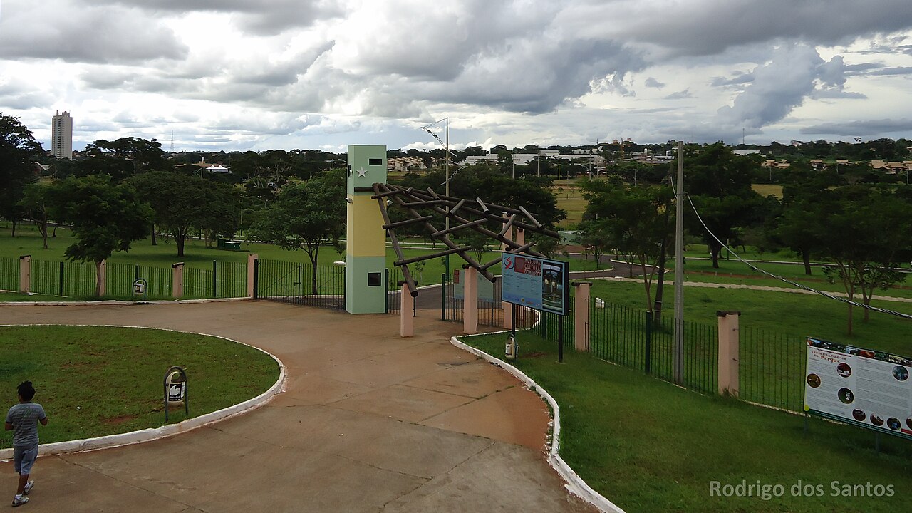 Entrada do Parque das Nações Indígenas com pórtico de troncos, torre verde e amarela, grades, caminhos, gramados e árvores sob céu nublado.